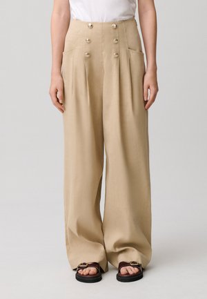 Pantaloni beige a gamba larga con vita alta, sei bottoni dorati, tasche laterali, indossati con top bianco e sandali marrone scuro.
