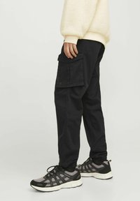 Black cargo pants made of durable material, featuring side pockets, a relaxed fit, and elastic cuffs, paired with black and grey sneakers.