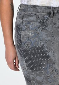 Jeans de mezclilla gris con un patrón floral texturizado, acentuados con pequeños tachuelas y un sutil tono azulado. Corte ajustado al cuerpo, vista posterior.