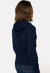 Haut à capuche tricoté bleu marine avec une texture douce, doté d'une poche au dos, de manches longues et d'une capuche avec cordon de serrage. Design simple et ajusté.