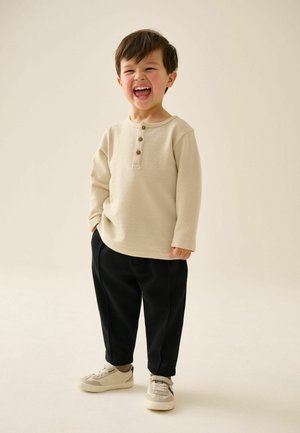 Jongen lacht, draagt een beige shirt met lange mouwen, zwarte broek en beige sneakers, staand tegen een effen lichte achtergrond.