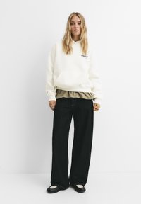 Överdimensionerad krämfärgad hoodie med framficka, lagd över en taupefärgad tröja; kombinerad med vida svarta jeans och svarta skor med vita detaljer.