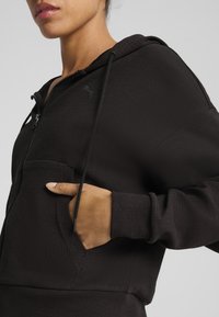 Black zip-up hoodie made of textured fabric, featuring a kangaroo pocket and adjustable drawstrings. Subtle branding on the chest.
