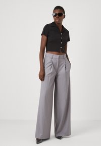 Chemise noire à boutons raccourcie associée à un pantalon large à rayures grises. Le mannequin porte des lunettes de soleil noires et des escarpins gris à bout pointu.