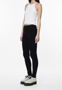 Mujer de pie vistiendo una blusa blanca sin mangas, pantalones skinny negros, calcetines blancos y zapatos de plataforma beige con cordones, contra un fondo blanco.