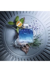 Bouteille de parfum en verre bleu avec un capuchon en argent, entourée de feuilles, de citrus, de lavande et d'éclaboussures d'eau sur une surface grise texturée.