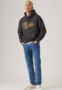 Joven con cabello rubio que lleva una sudadera oscura con texto amarillo, jeans azules y zapatillas blancas, de pie con las manos en los bolsillos.