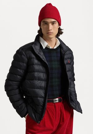Casaco puffer em tecido preto brilhante, combinado com um suéter xadrez em verde escuro e azul, calças de veludo cotelê vermelhas e um gorro de malha vermelho brilhante.