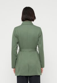 Προσαρμοσμένο πράσινο παλτό με ζώνη, που διαθέτει γιακά με σχισμή και μακριά μανίκια. Κατασκευασμένο από λείο ύφασμα με εφαρμοστή σιλουέτα.