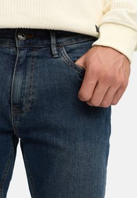 Hand in de voorzak van donkerblauwe spijkerbroek, gecombineerd met een roomkleurige, getextureerde trui zichtbaar bij de taille en mouw.