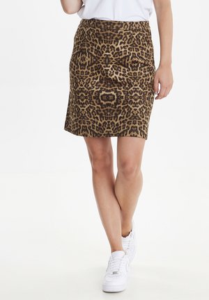Femme portant une jupe léopard mi-longue, des baskets blanches et un haut blanc, debout avec les jambes croisées sur fond blanc.
