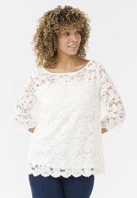 Top bianco in pizzo con maniche a tre quarti, motivo floreale, orlo a scalloped e scollatura ampia. Indossato sopra pantaloni in denim scuri.