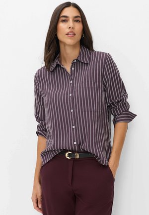 Femme portant une chemise rayée verticale violet foncé avec les manches retroussées et un pantalon assorti, debout avec une main dans la poche.
