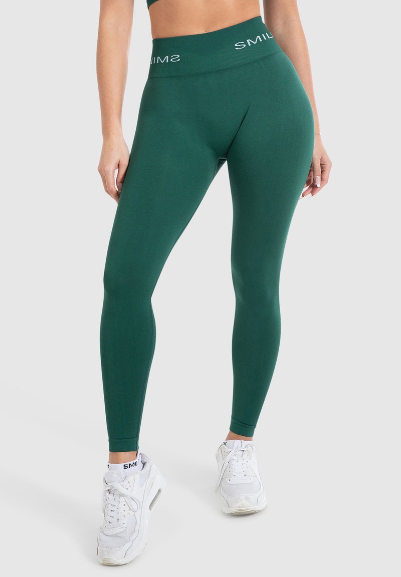 Groene leggings met een hoge tailleband met witte logotext, een gladde textuur en een aangesloten ontwerp, gecombineerd met witte sportschoenen.