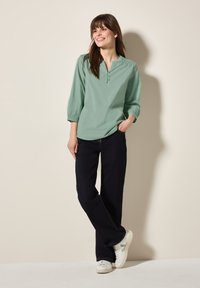 Blusa verde menta a maniche lunghe con bottoni, dettagli arricciati sulle spalle, abbinata a jeans a gamba larga blu scuro e sneakers bianche.