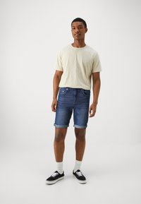 Ljusbeige kortärmad skjorta, denimshorts med uppvikta ärmar, mörkblå nyans, och svarta sneakers med vit sula och snören.