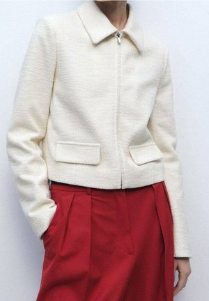 Personne portant une veste courte blanche texturée avec col et poches à rabat, associée à un pantalon rouge taille haute plissé.