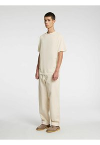 Beež lühikeste varrukatega t-särk koos lahtiste beežide pükstega ja heleroheliste kingadega. Kangas tundub olevat sile ja sellel on minimaalne disain.