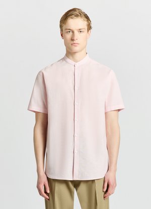 Junger Mann mit hellblonden Haaren, der ein blass rosa kurzärmeliges Hemd und beigen Hosen vor einem schlichten Hintergrund trägt.