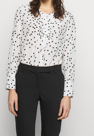 Femme portant un chemisier blanc à manches longues avec des volants et des pois noirs, rentrée dans un pantalon noir taille haute, debout devant un fond uni.