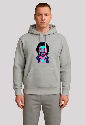 Mann trägt einen grauen Kapuzenpullover und eine Hose, der Kapuzenpullover zeigt ein buntes, stilisiertes Porträt eines lächelnden Mannes mit lockigem Haar und einem Bart.