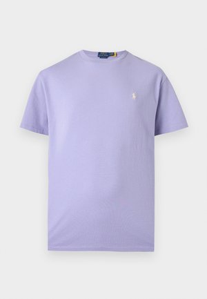 Homme à la tête rasée portant une chemise lavande à manches courtes, un pantalon blanc et des baskets beiges, debout avec une main dans la poche.