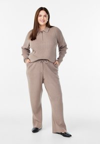 Bežs adīts džemperis ar rāvējslēdzēja apkakli un saskaņotiem šņorēšanas biksēm. Mīksta tekstūra un brīvs piegriezums, kombinēts ar melnām dzīvokļu kurpēm, kurām ir kniedes.