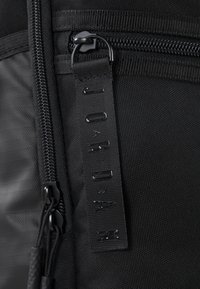 Fekete szövet hátizsák sima felülettel, dupla cipzárral és márkajelzéssel ellátott, dombornyomott feliratú címkével. A cipzárhúzók texturáltak a jobb fogás érdekében.