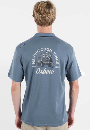 Homme portant un polo bleu avec un camion vintage et le texte "Having Good Times Since 1985 Oxbow" dans le dos.
