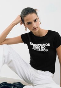 Schwarzes T-Shirt mit fettem weißen Text, der "ESCUCHAMOS PERO NO JUZGAMOS" liest. Das Modell trägt weiße Hosen und goldene Creolen. Einfarbiger Hintergrund.