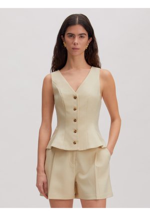 Vrouw met lang donker haar die een beige mouwloze peplumtop met knopen en bijpassende shorts draagt, staand met één hand in de zak.