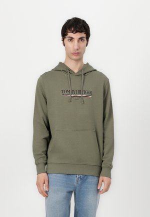 Olive green hoodie with a front pocket, drawstring hood, and printed logo. Features ribbed cuffs and hem. Worn with light blue jeans.