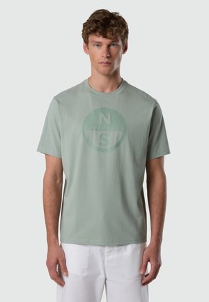Giovane uomo con capelli corti e ricci che indossa una maglietta verde chiaro con un logo circolare "N S North Sails" e pantaloni bianchi, in piedi in posizione neutra.