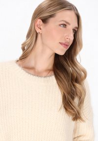 Kötött krémszínű pulóver, kerek nyakkivágással, finom szürke díszítéssel. A modellnek hosszú, hullámos haja van, és kis fülbevalót visel.