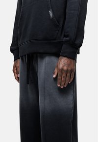 Zwarte hoodie met een voorzak, gecombineerd met grijze ombre joggingbroeken. Beschikt over een trekkoord in de taille en zichtbare stikdetails.