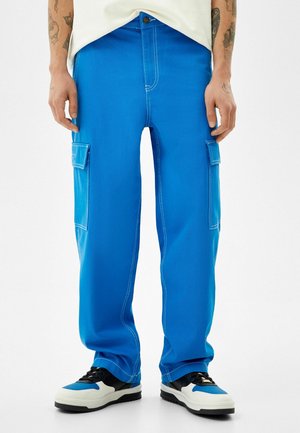 Mężczyzna w jaskrawo niebieskich spodniach typu cargo z białym przeszyciem oraz czarno-biało-niebieskich trampkach, stojący na białym tle.