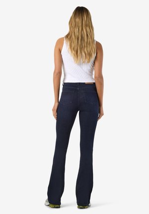 Vrouw met lang blond haar, een wit mouwloos topje en donkerblauwe uitlopende spijkerbroek, van achteren op een witte achtergrond.