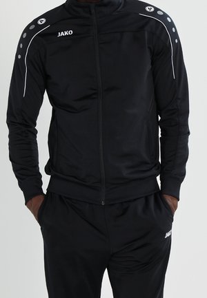 Zwart JAKO trainingspak met jas met ritssluiting en bijpassende broek, met wit logo en decoratieve stippen op de schouders, gedragen met handen in de zakken.