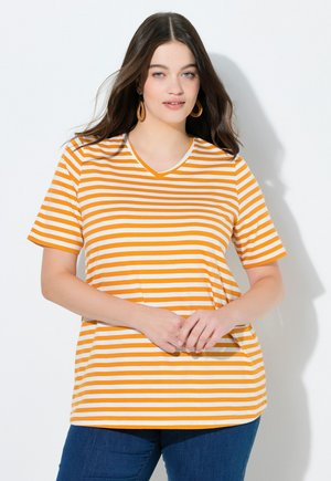 Frau mit langen dunklen Haaren, die ein orange-weiß gestreiftes T-Shirt mit kurzen Ärmeln und blaue Jeans trägt, steht vor einem weißen Hintergrund.