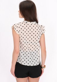 Doorzichtige blouse met korte mouwen in crème met zwarte stippen. Kenmerkt rimpelige mouwen en een ronde zoom, gecombineerd met zwarte shorts.