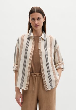 Kobieta o długich brązowych włosach, ubrana w beżowo-czarną koszulę w paski na guziki, na brązowym topie i brązowych spodniach z sznurkiem w pasie.