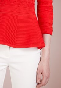 Haut à manches longues rouge texturé et pantalon blanc avec une main droite visible sur un fond clair uni.