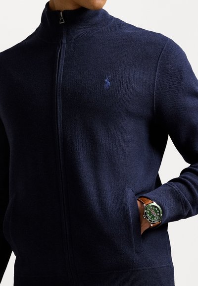 Cardigan cu fermoar de culoare bleumarin, cu guler înalt, fabricat dintr-un material tricotat texturat. Dispune de un mic logo și o buzunar frontal. Ceas marron vizibil.