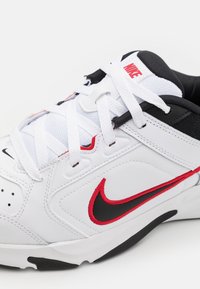 Nike sneaker in white leather featuring black and red accents, round toe, textured overlays, and white laces. Design includes logo detail.