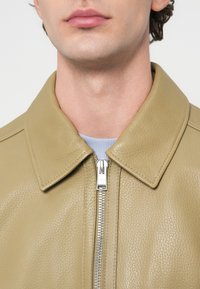 Κοντινό πλάνο ενός ατόμου που φοράει μπεζ δερμάτινο μπουφάν με υφή, με ασημί φερμουάρ και ανοιχτό μπλε πουκάμισο από κάτω.