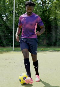 Camisola roxa e rosa da Puma, calções pretos e meias até ao joelho. Jogador dribla uma bola de futebol azul e amarela num campo de relva.