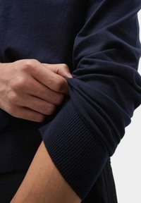 Námořnický pletený svetr s žebrovaným manžetou. Ruka drží rukáv, což ukazuje na měkkou strukturu a volný střih látky.