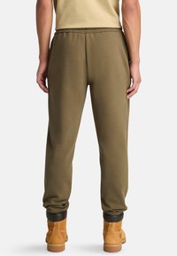 Eine Person steht mit dem Rücken zum Betrachter, trägt olivgrüne Jogginghosen und helle Arbeitsschuhe und zeigt einen entspannten, lässigen Stil.