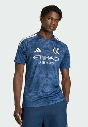 Mann trägt blaues NYCFC-Fußballtrikot mit geometrischem Muster und Logos von Etihad Airways und Adidas, steht mit einer Hand in der Hosentasche.