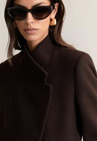 Mujer con abrigo oscuro de gran tamaño, gafas de sol negras en forma de ojo de gato y grandes pendientes de aro dorados, con el cabello castaño liso.
